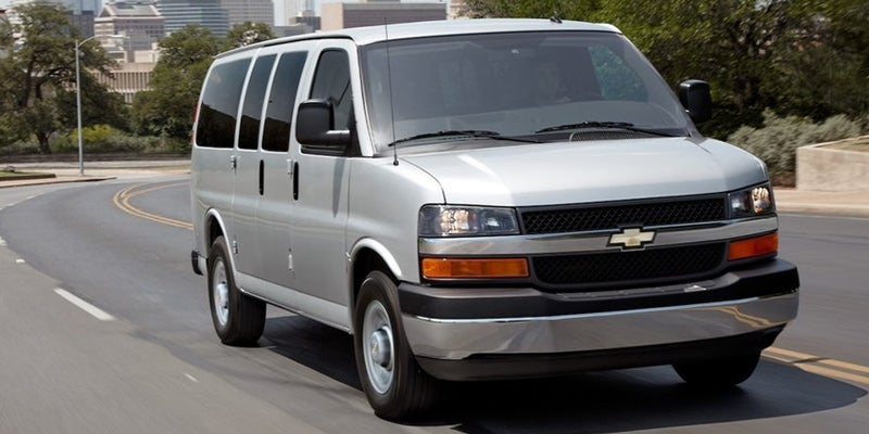 Silver Chevrolet Express passenger van driving on a curved roadway, featuring a chrome Chevrolet grille