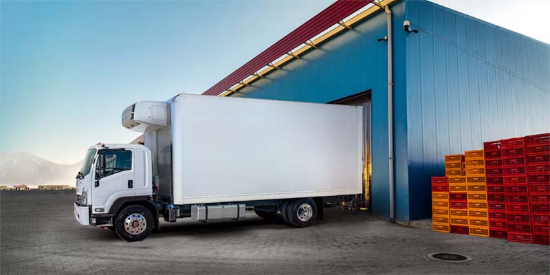 White box truck positioned at a loading dock outside a warehouse, featuring an enclosed cargo body designed for transporting goods.