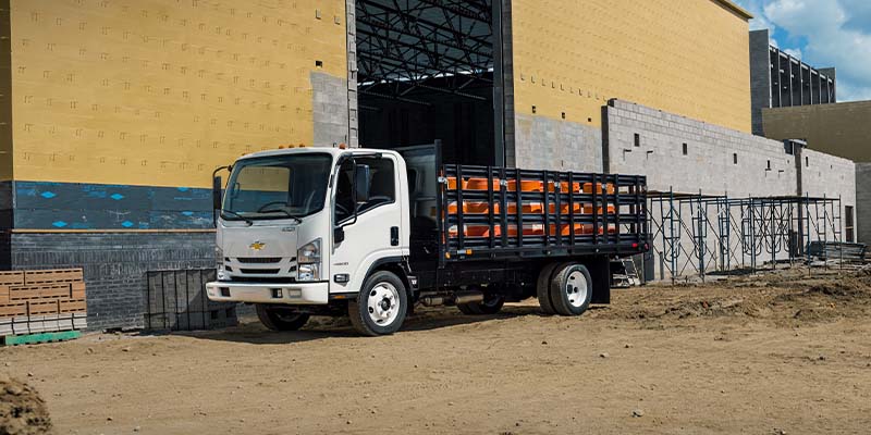 White medium-duty stake bed truck parked at a construction site, featuring side rails for carrying building materials