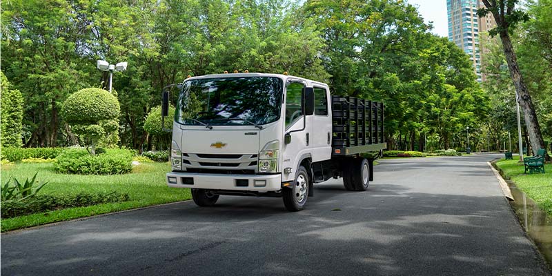 White Chevrolet medium-duty stake bed truck driving on a tree-lined road, featuring a crew cab