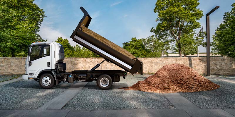 White medium-duty dump truck unloading a raised dump bed filled with mulch in an outdoor commercial yard surrounded by trees and a stone wall