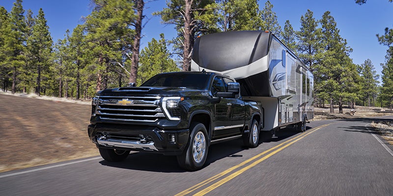 Black Chevrolet Silverado HD pickup truck towing a large travel trailer on a paved road through a forested landscape with tall pine trees
