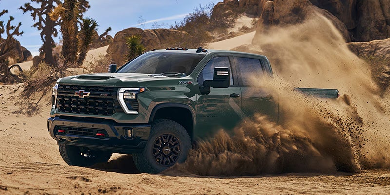 Green Chevrolet Silverado HD ZR2 pickup truck driving off-road through a desert landscape, kicking up a large cloud of sand and dust around the tires.