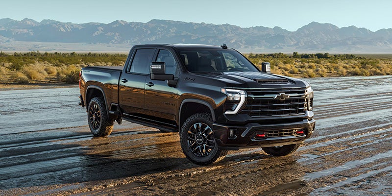 Dark-colored Chevrolet Silverado pickup truck parked on a wet dirt road with mountains and open terrain visible in the background.