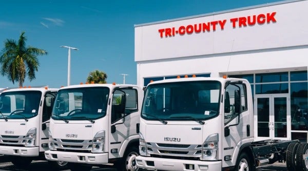 White Isuzu cab-chassis trucks parked in front of a TRI-COUNTY TRUCK dealership under a blue sky.