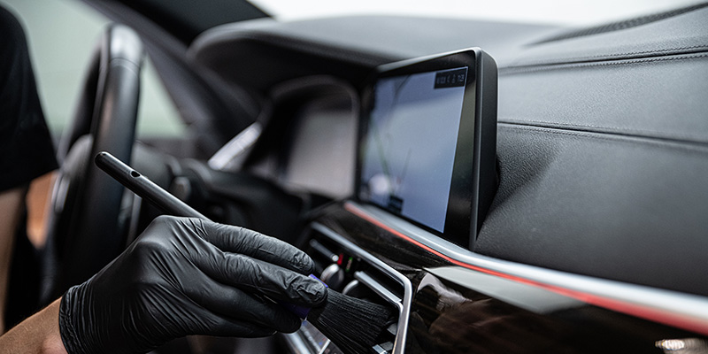 Hand in black glove cleaning car interior with a detailing brush near the infotainment screen and vents