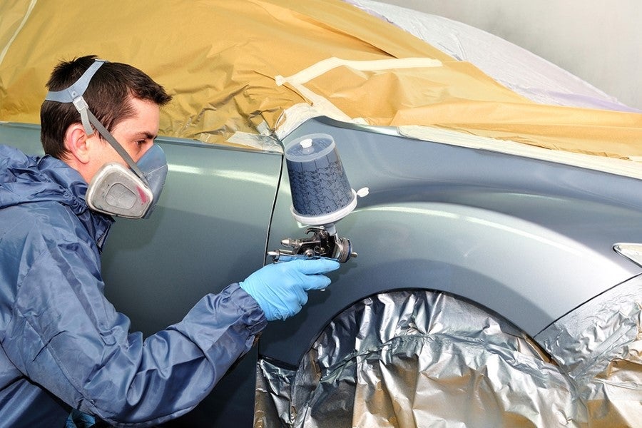 Man in respirator and blue suit spray painting a car light blue, with parts masked off