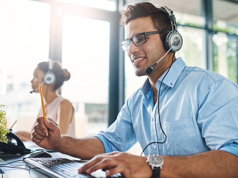 Smiling man in glasses and headset working at computer, holding a pencil. Another person works in background