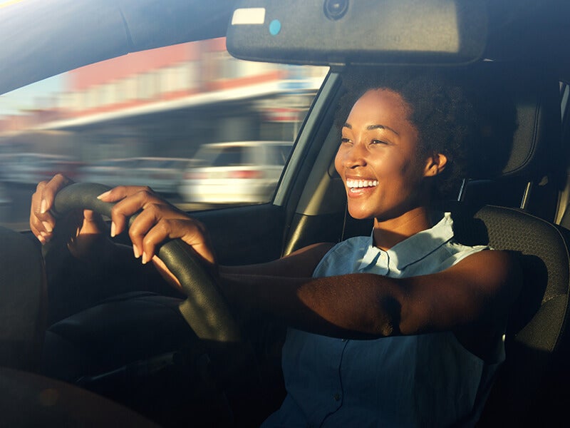 Smiling Black woman happily driving a car with her hands on the steering wheel, sunlight on her face
