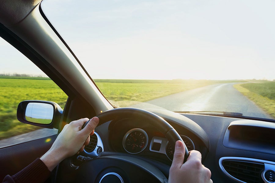 Hands on a steering wheel, driving on a winding country road through green fields under a bright, sunny sky