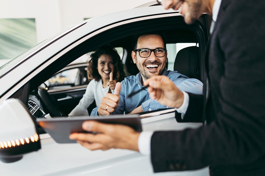 Happy couple in a new white car giving thumbs up to a salesperson holding a tablet