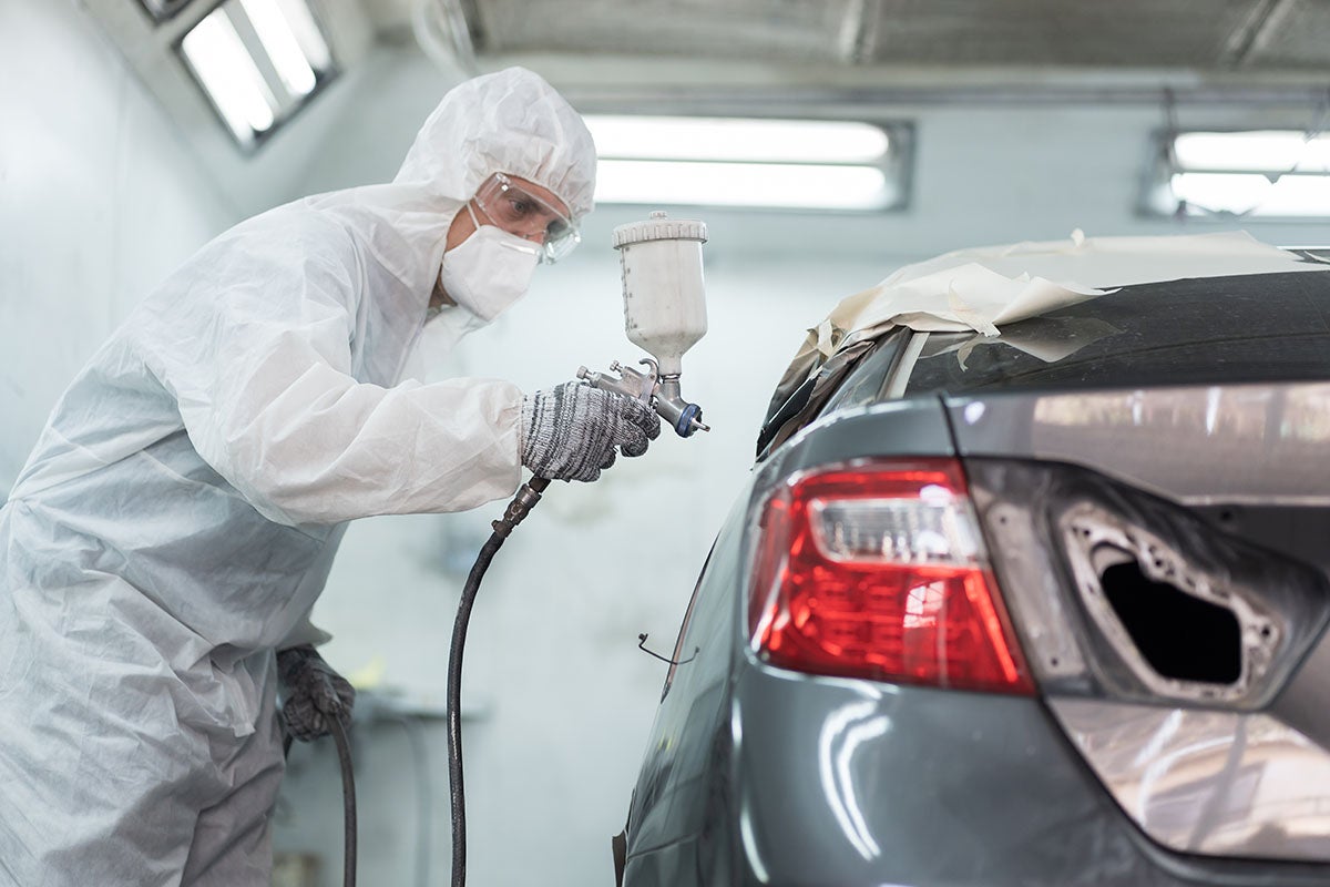 Person in protective suit and mask spray painting the rear of a grey car in a garage