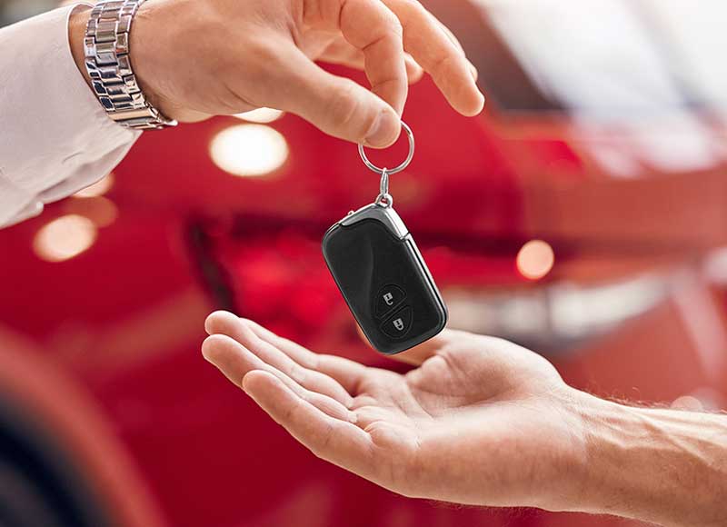 A hand holding a car key fob is passing it to another open hand, with a red car in the background.