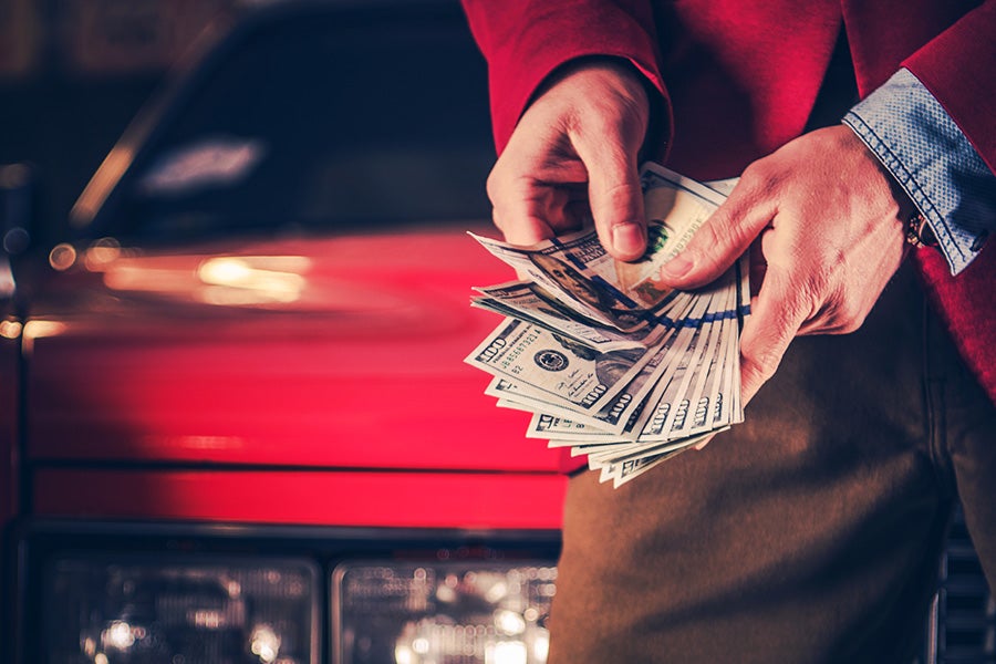 Hands holding a large stack of hundred-dollar bills in front of a blurry red car.